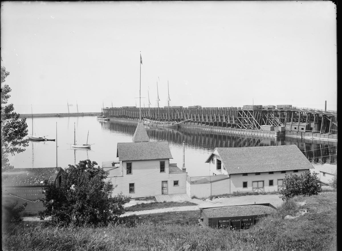 Coal and ore docks at Oswego around 1900: a long timber trestle stacked with loaded rail cars reaching across the harbour, schooner masts behind it
